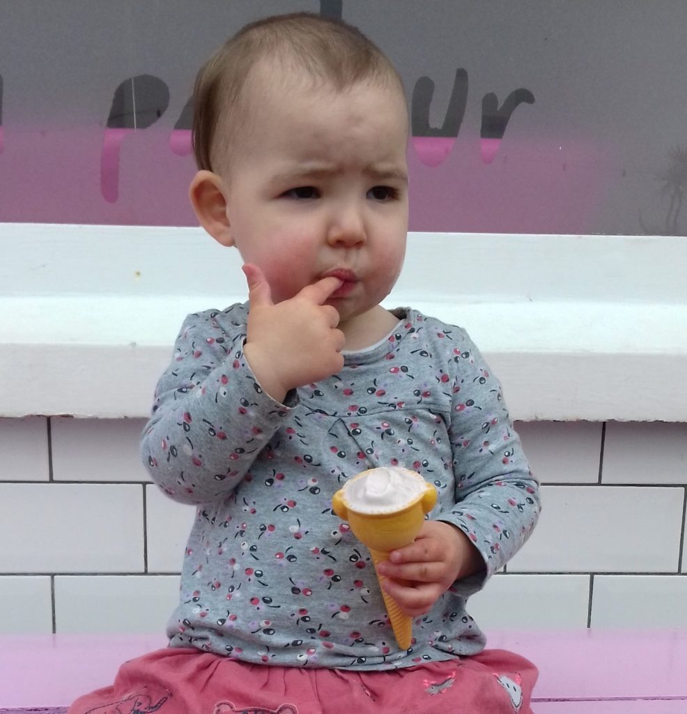 photograph of a toddler girl holding an ice cream in one hand and licking her finger on the other hand, outside an ice cream shop